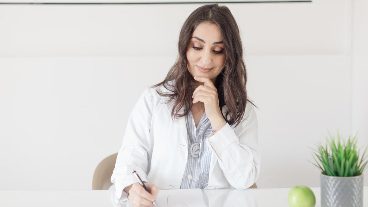 A female nutritionist in a lab coat writing notes with an apple and plant on the desk.